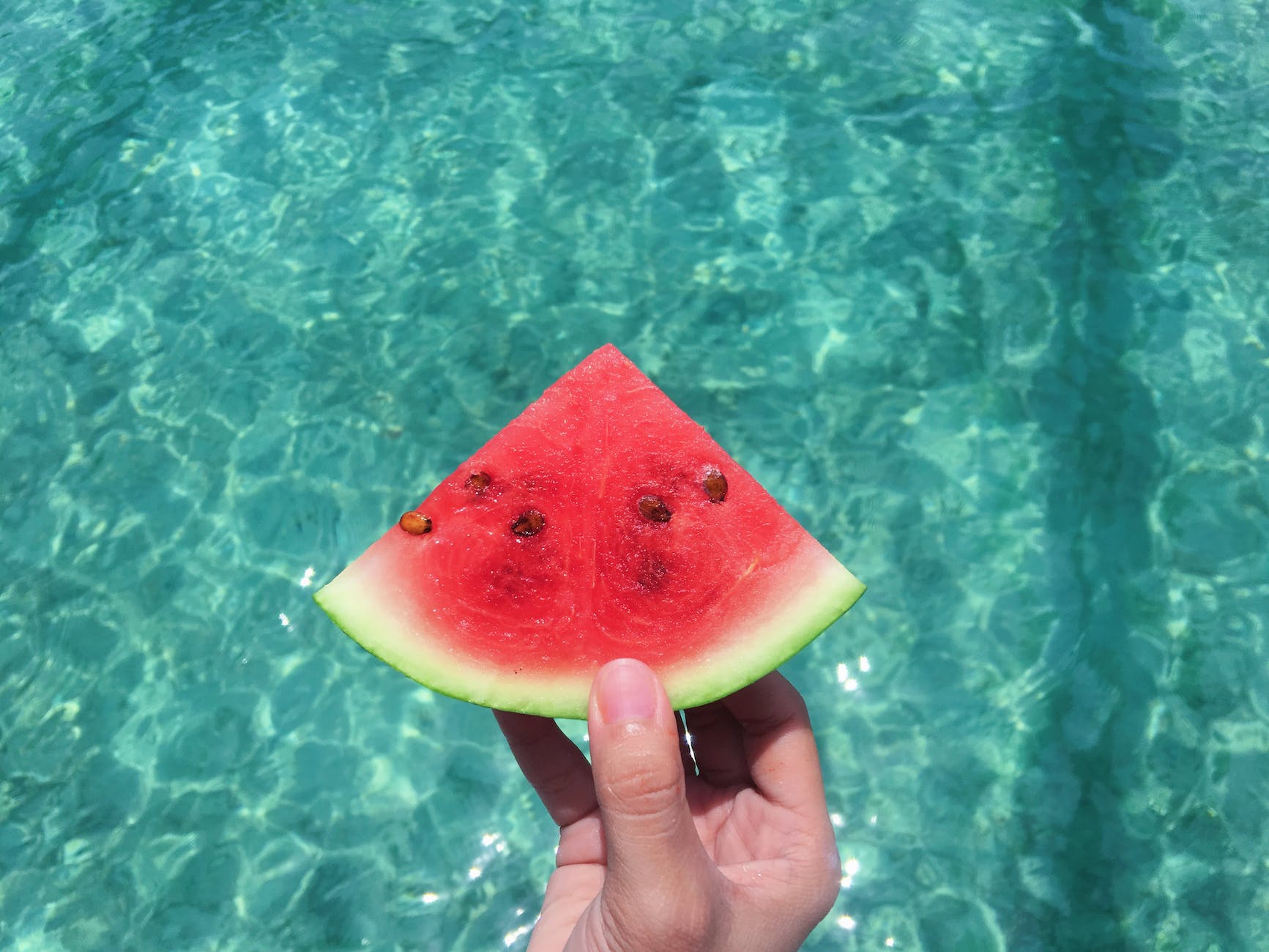 hand holding a slice of watermelon with blue swimming pool water in the background melone parfum